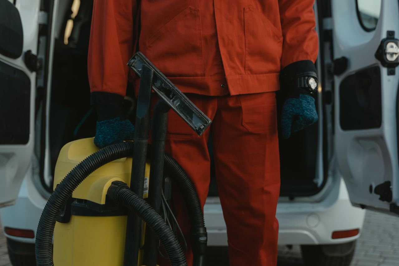 A cleaner in a red uniform stands beside a van with a vacuum, prepared to provide service.