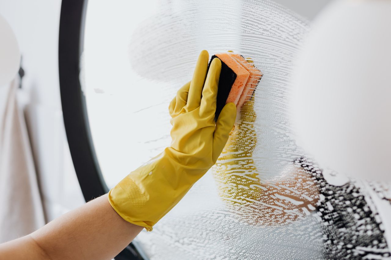 about-us Personne anonyme portant des gants en caoutchouc jaunes et utilisant une simple éponge pour nettoyer le miroir de la salle de bain
